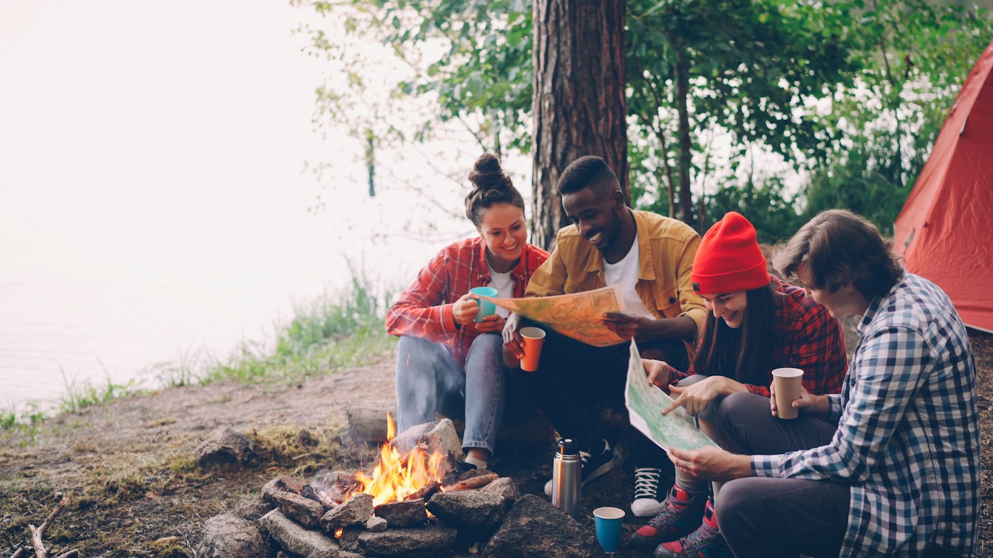Four friends review maps around a campfire by the water.